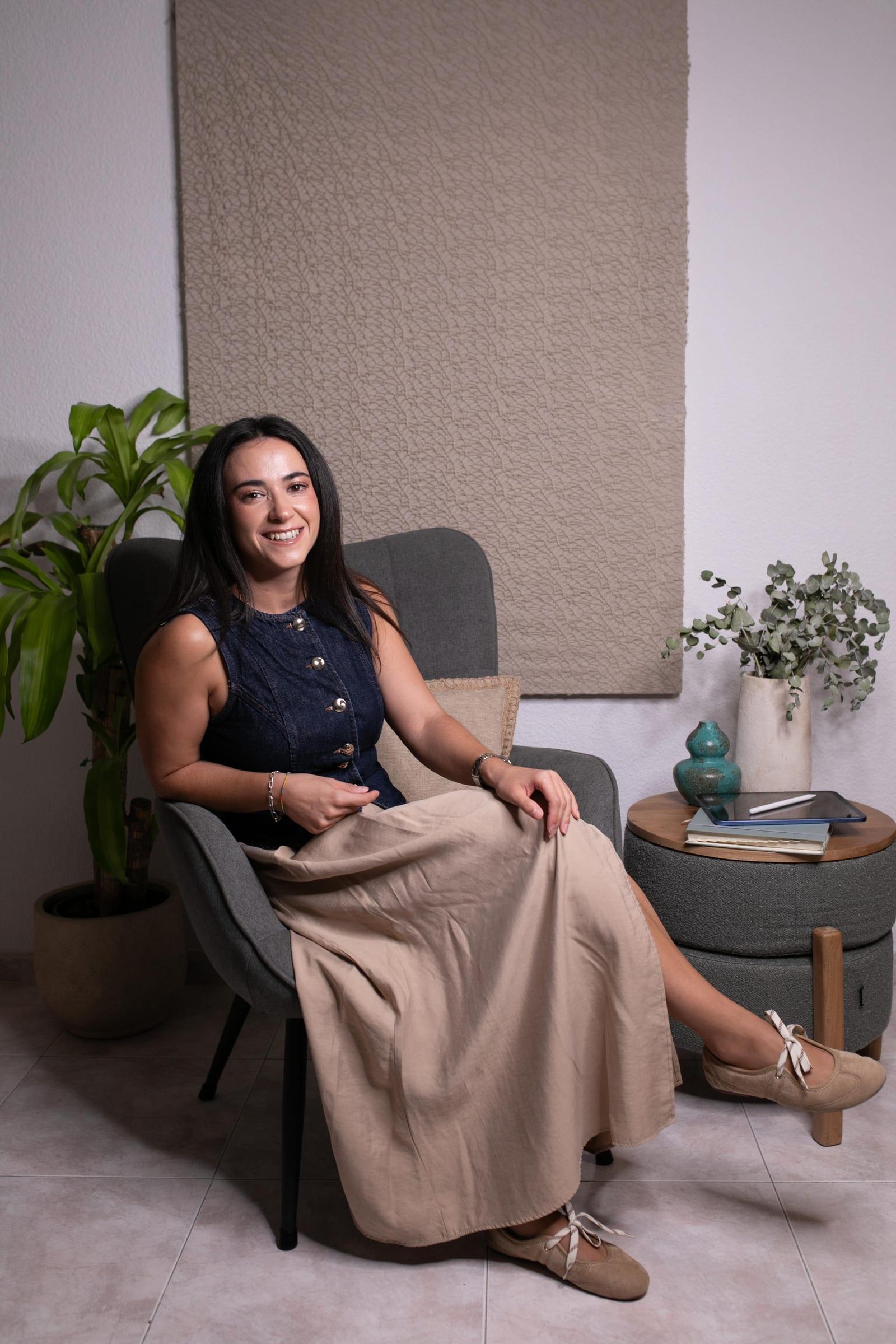 Elena Sánchez Carrasco sentada en un sillón gris en su consulta, junto a una planta y una lámpara